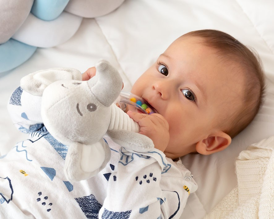Bebé a morder um brinquedo de elefante de peluche cinzento, vestido com macacão branco com padrões azuis e pretos.
