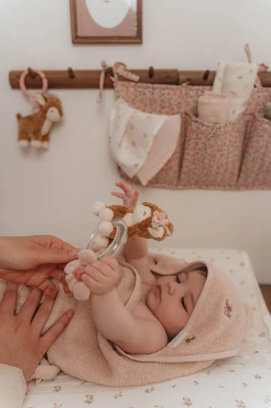 Bebé enrolado numa toalha rosa com brinquedo de chocalho em ambiente decorado.