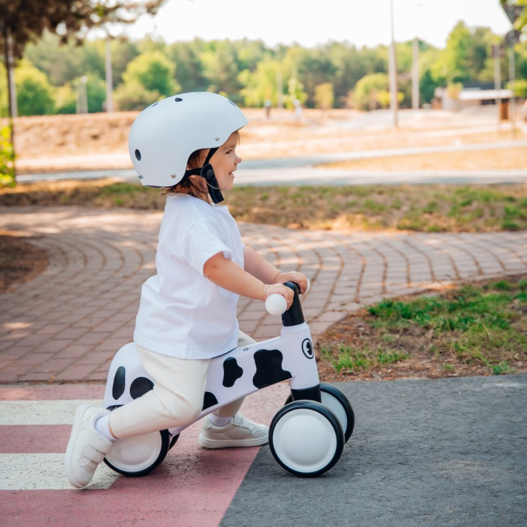 Criança pequena com capacete branco numa bicicleta de equilíbrio com padrão branco e preto em ciclovia urbana