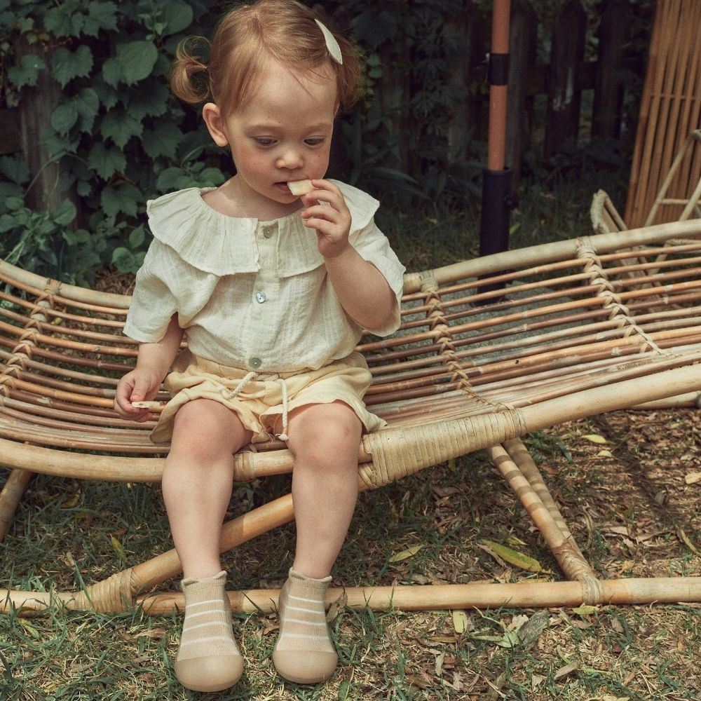 Criança sentada num banco de vime com roupa clara e meias bege.