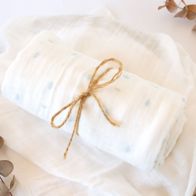 Fralda branca fina com padrão azul enrolada com cordão de juta numa cesta de vime com tecido branco e folhas secas