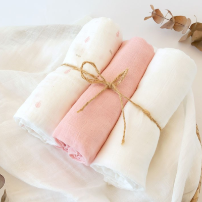 Três panos de tecidos enrolados, branco, rosa claro e branco com padrão, sobre pano branco maior com folhas secas.