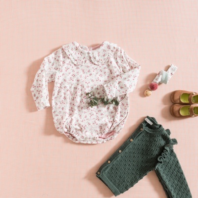 Conjunto de roupa de bebé com body floral branco, calças verdes de malha, sapatos castanhos e cinto colorido sobre fundo rosa xadrez