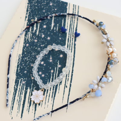 Pulseira de contas translúcidas e fio de cabelo decorado com flores brancas e cristais azuis sobre fundo branco e azul