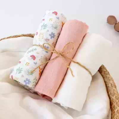 Cesta de vime com três toalhas enroladas de algodão ou linho, cores branco, rosa e floral, fundo branco minimalista