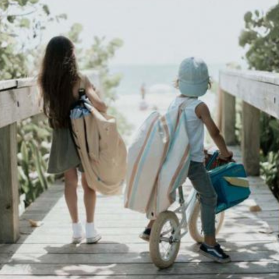Duas crianças numa passadeira de madeira junto à praia, uma com cadeira de praia enrolada e outra numa bicicleta sem pedais azul