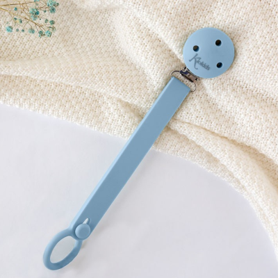 Prendedor de chupeta azul claro com fecho redondo e texto Kiza, sobre manta bege