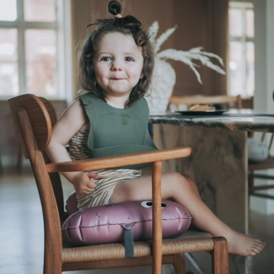 Criança pequena sentada numa cadeira de madeira com almofada roxa e babete verde.
