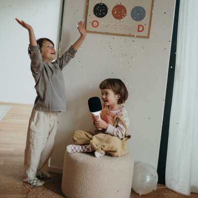 Duas crianças em divisão com pufe bege, microfone preto com luz, quadro colorido e cortina branca.