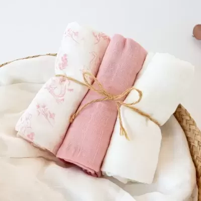 Três panos de tecido macio, um estampado rosa, outro rosa liso e outro branco, enrolados e amarrados com corda numa cesta de vime.