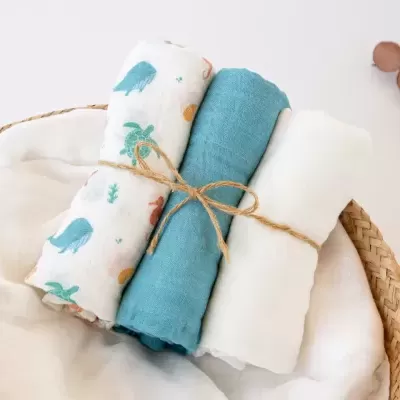 Três toalhas enroladas, uma com padrão de animais marinhos, uma azul e uma branca, amarradas com cordão de juta num cesto de verga.