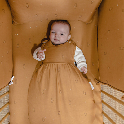 Bebé num saco de dormir castanho claro com padrão de urso dentro de cama de grades com protetor almofadado com o mesmo padrão