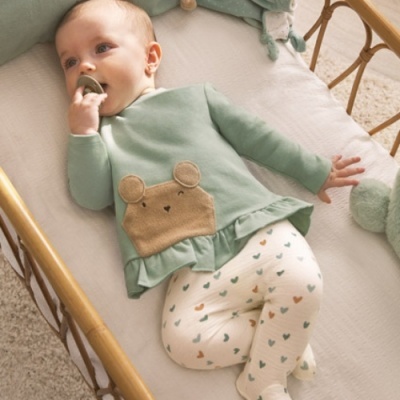 Bebé com roupa verde água e calças brancas com corações num berço de madeira.