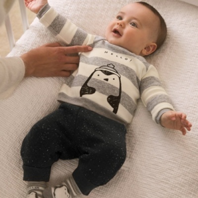 Bebé com camisola de pinguim e calças pretas deitado numa superfície clara