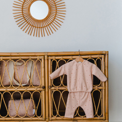Conjunto de roupa de bebé cor-de-rosa com pequenos corações pendurado em móvel de rattan com almofadas cor-de-rosa e espelho redondo em rattan na parede.