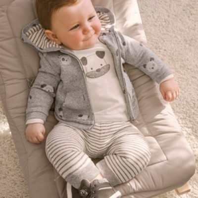 Bebé com roupa cinzenta e branca sentado num assento acolchoado bege