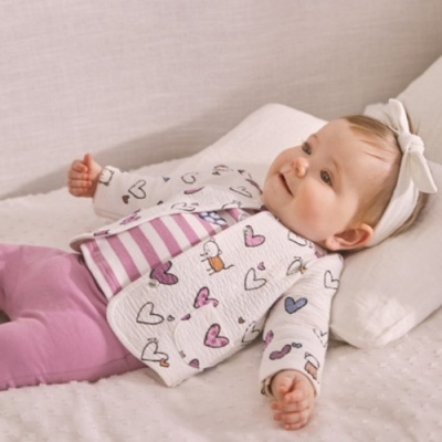 Bebé vestindo casaco acolchoado com corações, t-shirt às riscas, calças cor-de-rosa e faixa branca com laço deitada num colchão branco