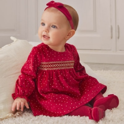 Bebé com vestido vermelho de padrões brancos e faixa vermelha no cabelo sentada num tapete branco.