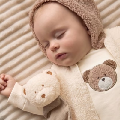 Bebé adormecido com roupa de urso bege e castanha, chapéu de pelo e brinquedo de urso