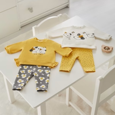 Dois conjuntos infantis em mesa branca, um amarelo e cinzento com flores, outro branco e amarelo com pontos e ursos desenhados.