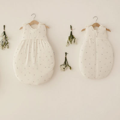 Dois sacos de dormir de bebé branco com padrões cinza pendurados em cabides numa parede clara com flores secas.