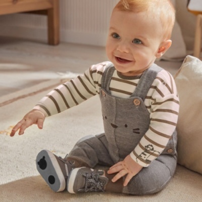 Bebé vestido com macacão cinza e camisa às riscas, sentado no chão interior