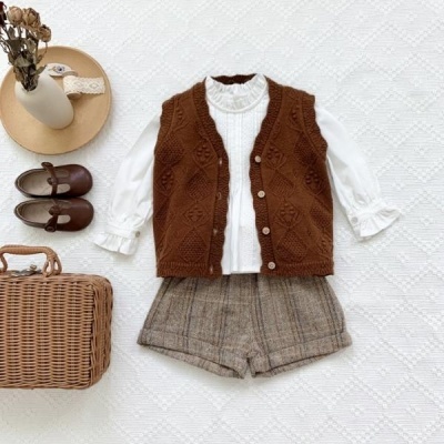 Conjunto infantil com colete malha castanho, camisa branca, calções xadrez, sapatos castanhos e mala de palha sobre fundo branco