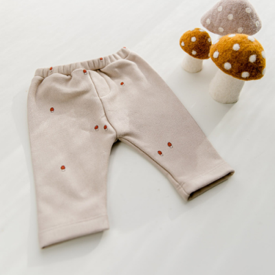 Calças de criança bege claro com pequenas bolinhas castanhas em tecido macio junto a três cogumelos decorativos em feltro.