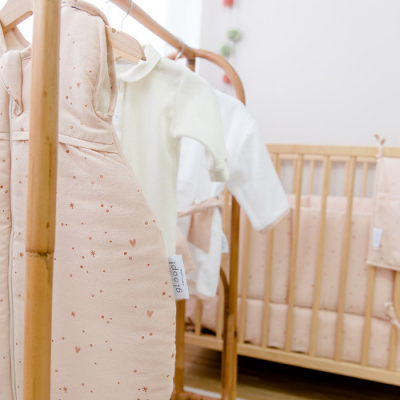 Roupa de bebé rosa claro com padrões e roupas brancas junto a berço de madeira clara em ambiente nobre