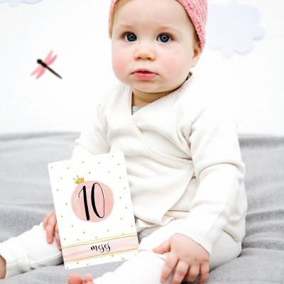 Bebé com fato branco e gorro rosa segurando cartão 10 meses sentado sobre superfície cinzenta