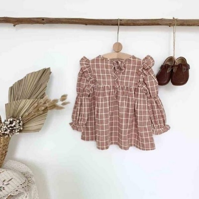 Vestido xadrez castanho e branco de criança com mangas compridas pendurado em cabide ao lado de sapatos castanhos.