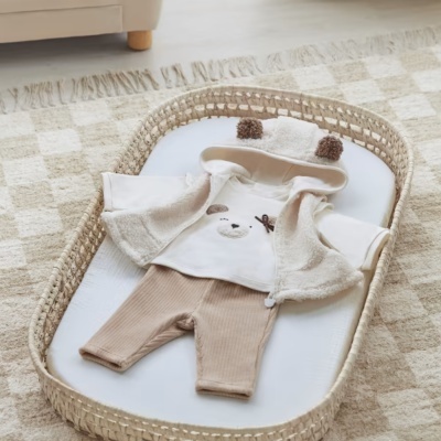 Roupa de bebé composta por camisola branca com capuz com orelhas castanhas, casaco aberto bege claro e calças castanhas de malha numa cesta de vime sobre tapete quadriculado.