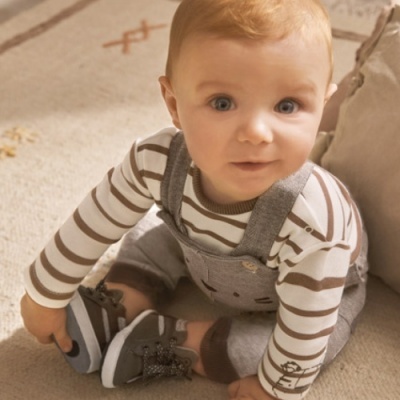 Bebé sentado com camisola às riscas, jardineiras cinzentas e sapatinhos cinzentos