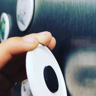 Íman redondo branco com disco preto sendo segurado por uma mão junto a superfície metálica magnética