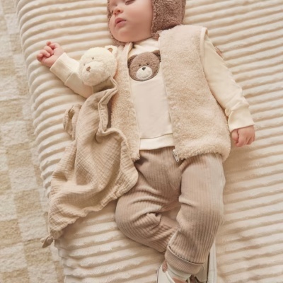 Bebé vestido com roupas de tecido felpudo e calças de risca fina, deitado numa manta bege ondulada