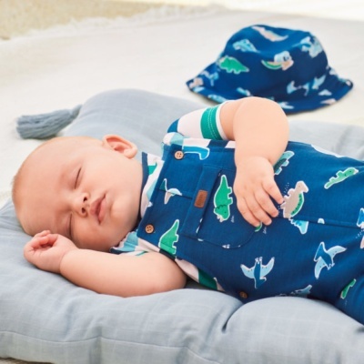 Bebé a dormir com jardineira azul de dinossauros e boné azul sobre almofada cinzenta