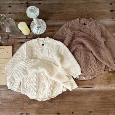 Conjuntos de roupa de bebé em malha creme e castanha clara com padrões texturizados sobre mesa de madeira com castiçal e vela