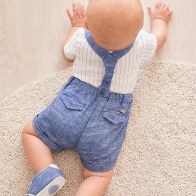 Bebé debruçado usando body branco com riscas azuis e calção azul com bolsos sobre tapete bege.
