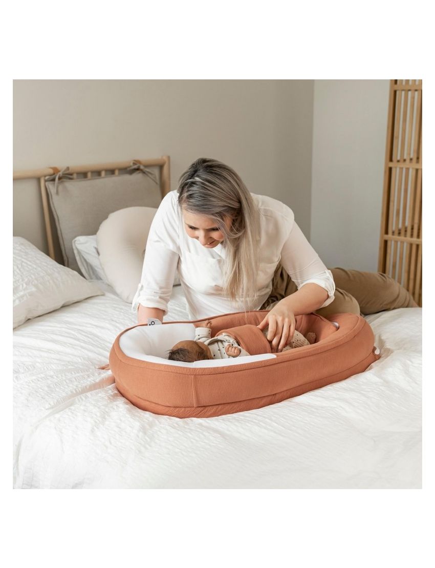 Berço portátil castanho claro para bebés numa cama branca com mulher a cuidar