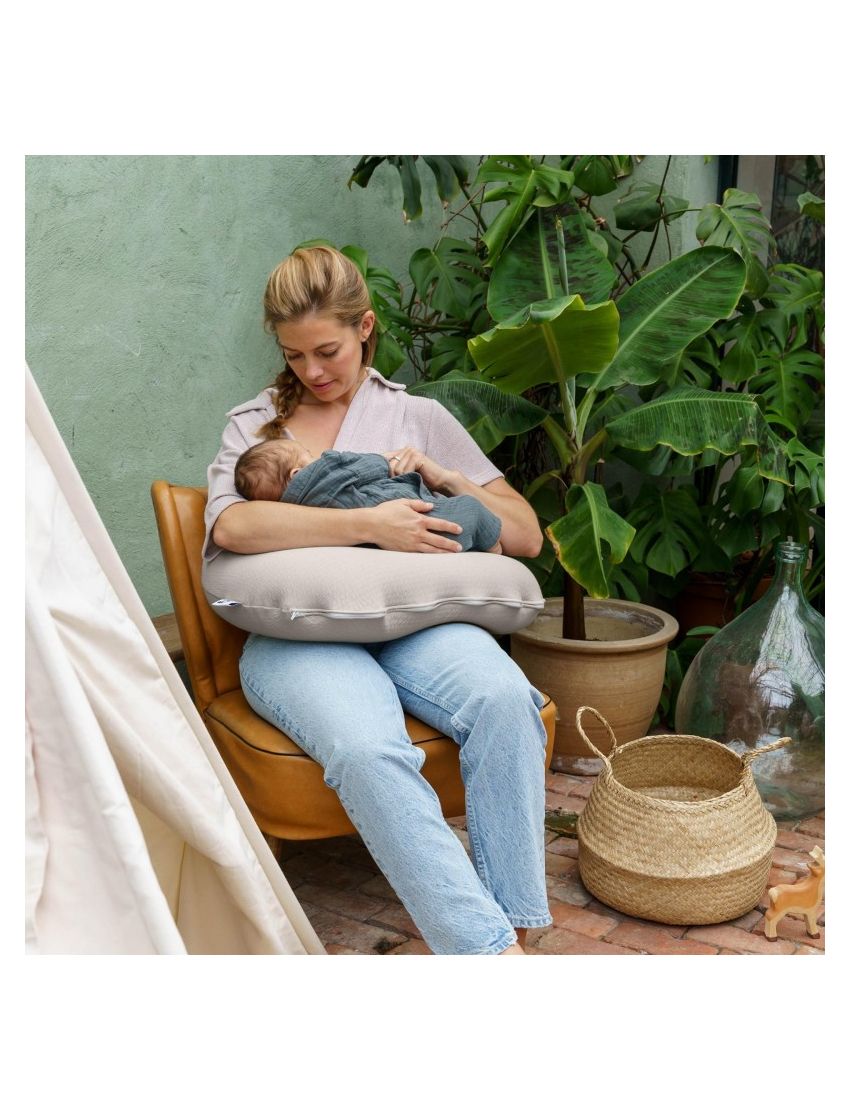 Mulher amamenta bebé com almofada, cadeira castanha, plantas e objetos em pátio
