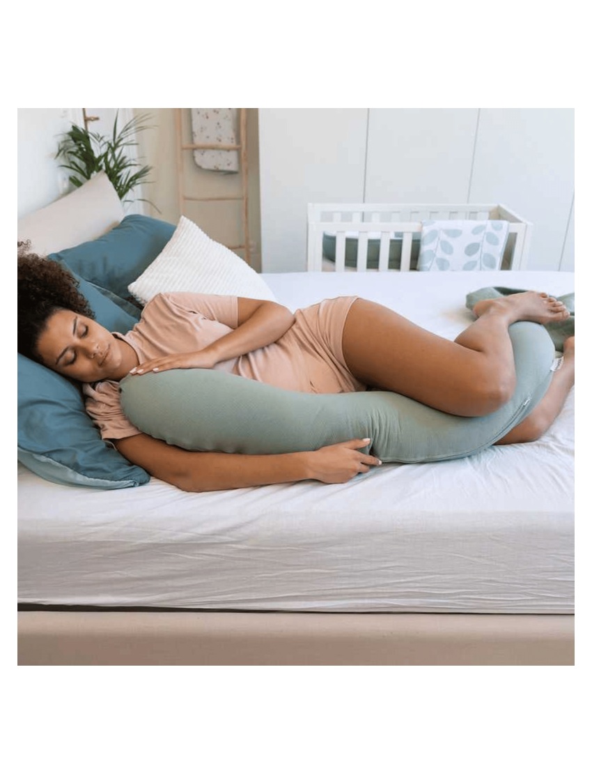 Mulher a dormir abraçada a travesseiro de corpo verde claro numa cama com almofadas e berço ao fundo.