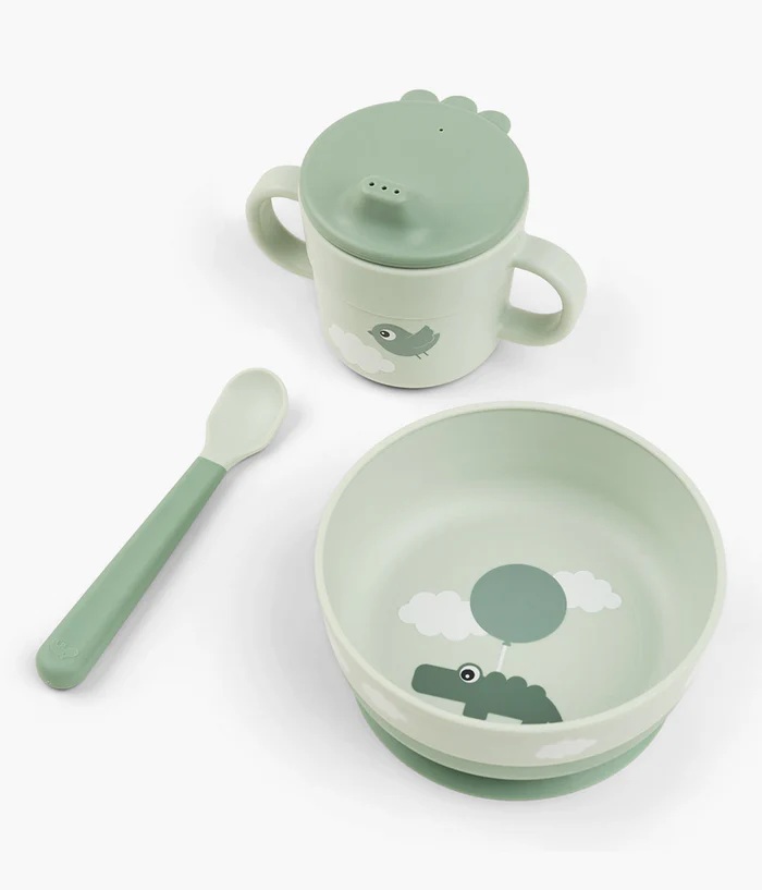 Conjunto infantil com caneca verde clara com tampa e bico, colher verde e taça verde com desenho de crocodilo e balão.
