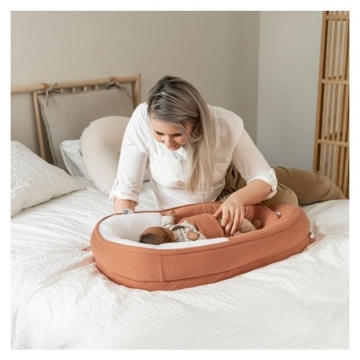 Berço portátil castanho claro para bebés numa cama branca com mulher a cuidar