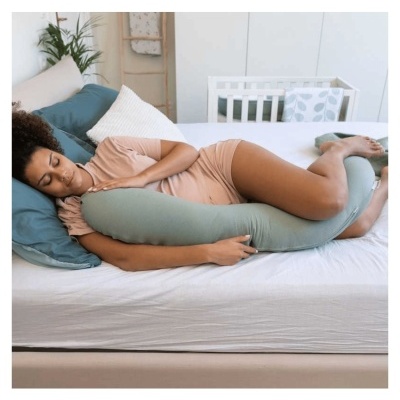 Mulher a dormir abraçada a travesseiro de corpo verde claro numa cama com almofadas e berço ao fundo.