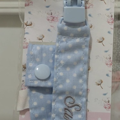 Clip para chupeta azul claro com bolinhas brancas e nome bordado Santiago em embalagem rosa decorada.