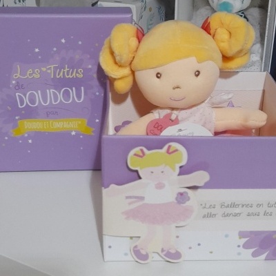 Boneca de pelúcia loira em caixa roxa com texto Les TutuS de DOUDOU