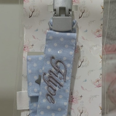 Prende-chuchas azul claro com bolinhas brancas e texto bordado 'Aline' em embalagem rosa com flores