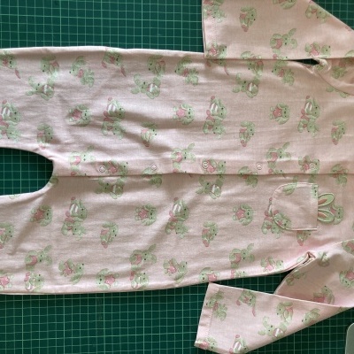 Pijama cor-de-rosa com padrão de coelhos verdes e bolso com orelhas bordadas, sobre base verde quadriculada.
