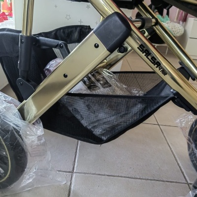 Carrinho de bebé com estrutura dourada e preta, rodas com plástico protetor e cestinha inferior preta