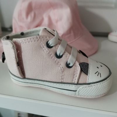 Ténis de bebê rosa com desenho de gato na biqueira, exibido sobre superfície branca